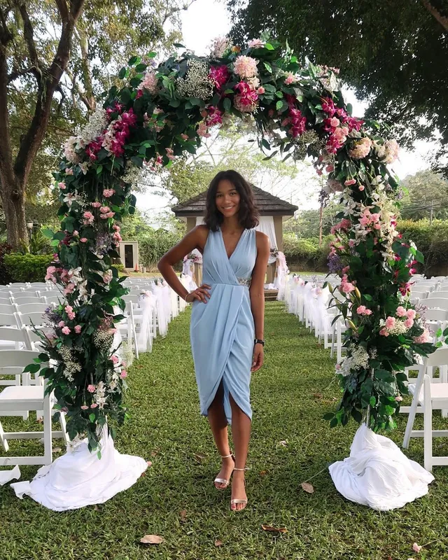 Greta Onieogou wearing a blue wrap dress and pale heels beneath a floral arch.