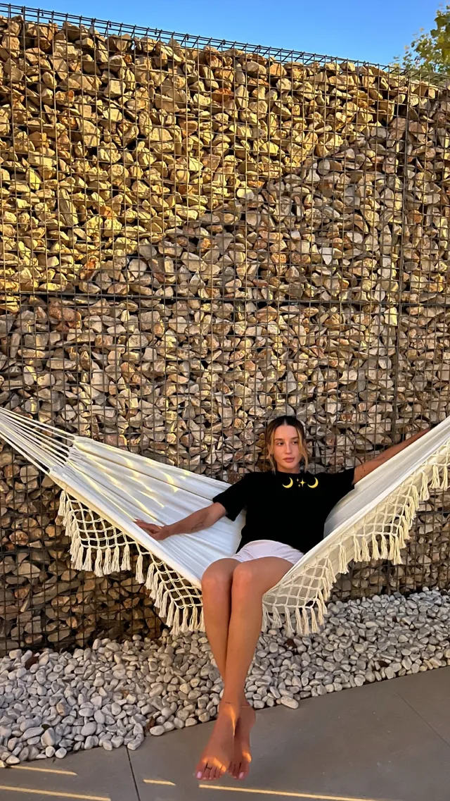María Pedraza sitting barefoot in a hammock in a dark top and light shorts.