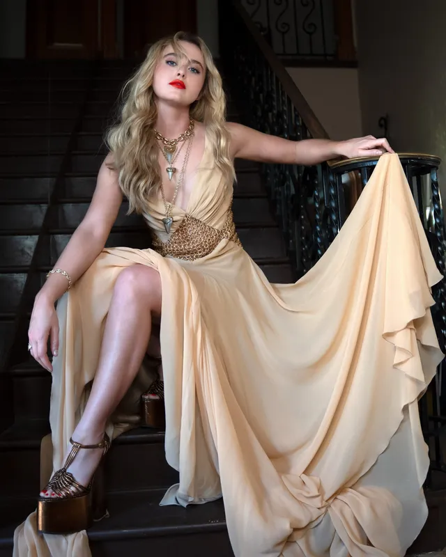 Kathryn Newton posing on a staircase in a gold gown and heels.