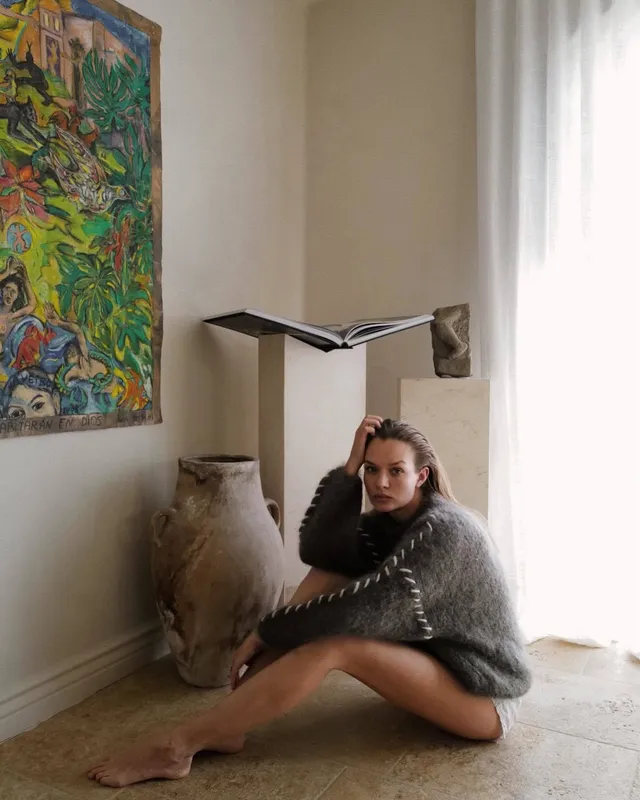 Josephine Skriver sitting barefoot on a rug in a dark knit outfit.