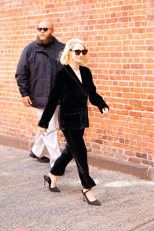 Jessica Chastain walking beside a brick wall in black pumps.