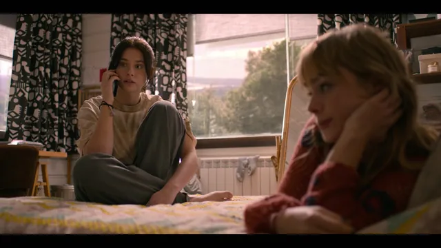 Nicole Wallace sitting barefoot on a bed beside another woman in a scene.