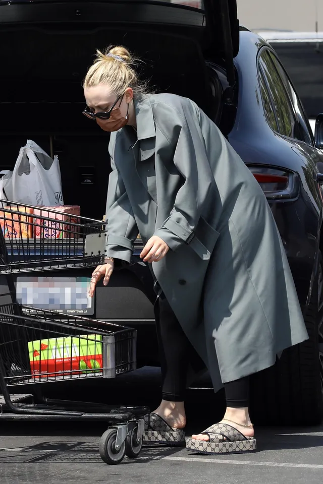 Dakota Fanning standing beside a car in a gray coat and patterned sandals.