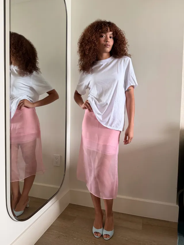 Whitney Peak posing indoors in a white T-shirt, pink skirt and pale heels.
