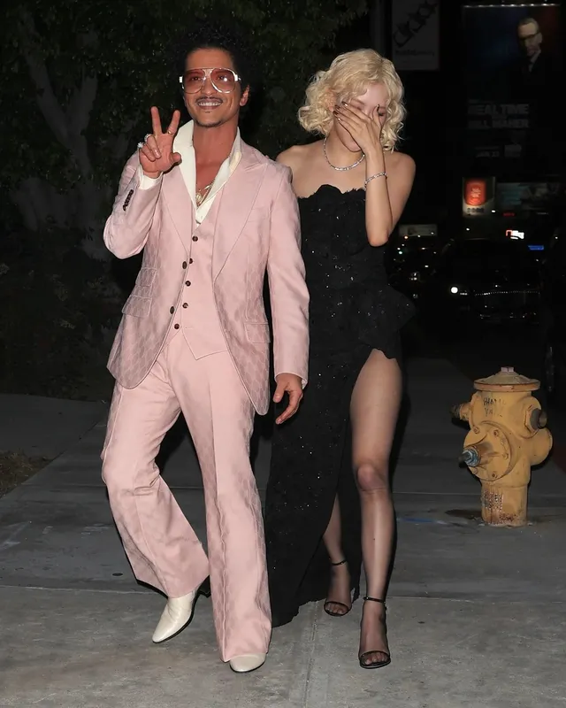Rosé walking outside at night in a black gown, sheer tights and ankle-strap heels.
