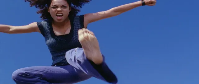 Rosario Dawson posing against a blue background with one foot raised.
