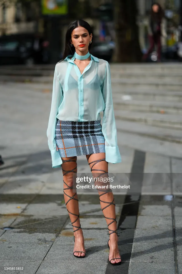 Bruna Marquezine wearing a pale blouse, checked skirt and black lace-up heels on a street.