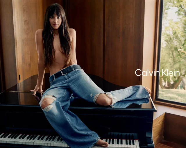 Dakota Johnson posing barefoot on a piano in jeans.