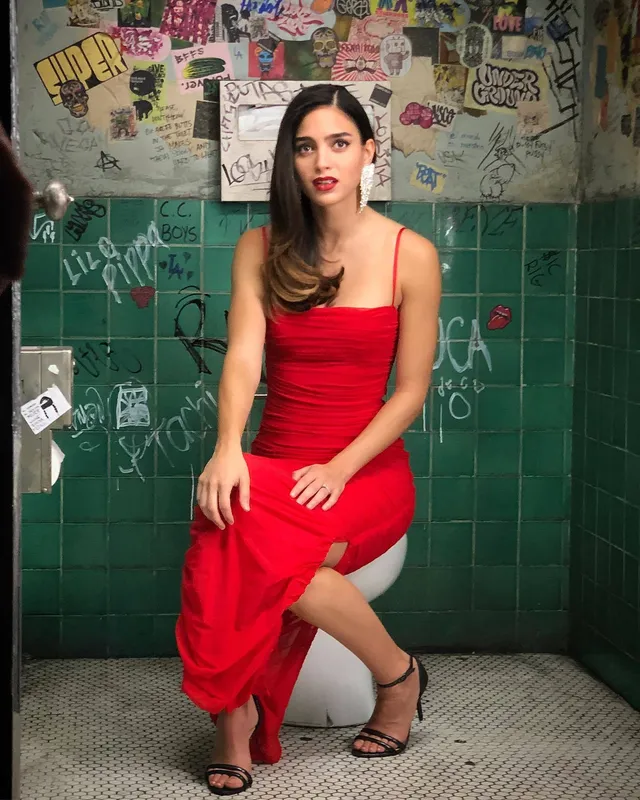 Melissa Barrera sitting in a red dress and black heeled sandals.