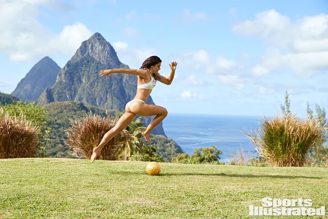 Alex Morgan running barefoot on grass beside a football during a swimwear shoot.