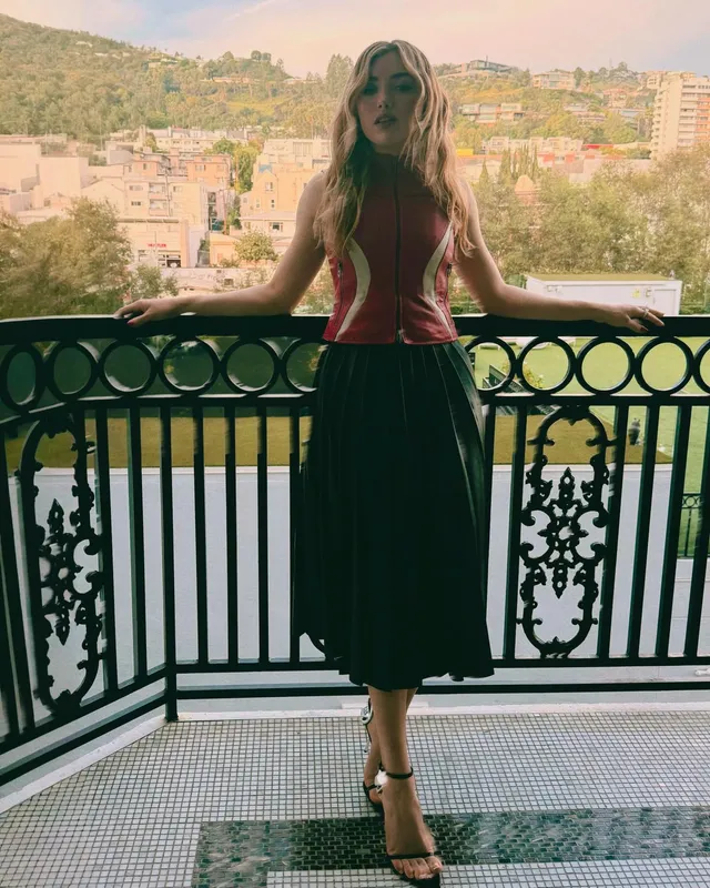 Peyton List wearing a red top and black heels on a balcony.