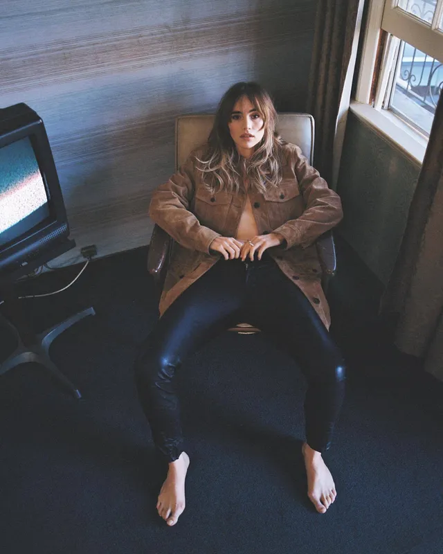 Suki Waterhouse sitting in an armchair in a brown jacket and jeans.