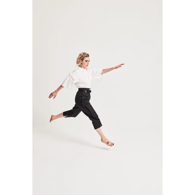 Vanessa Kirby a studio portrait leaping in dark trousers and a white shirt.