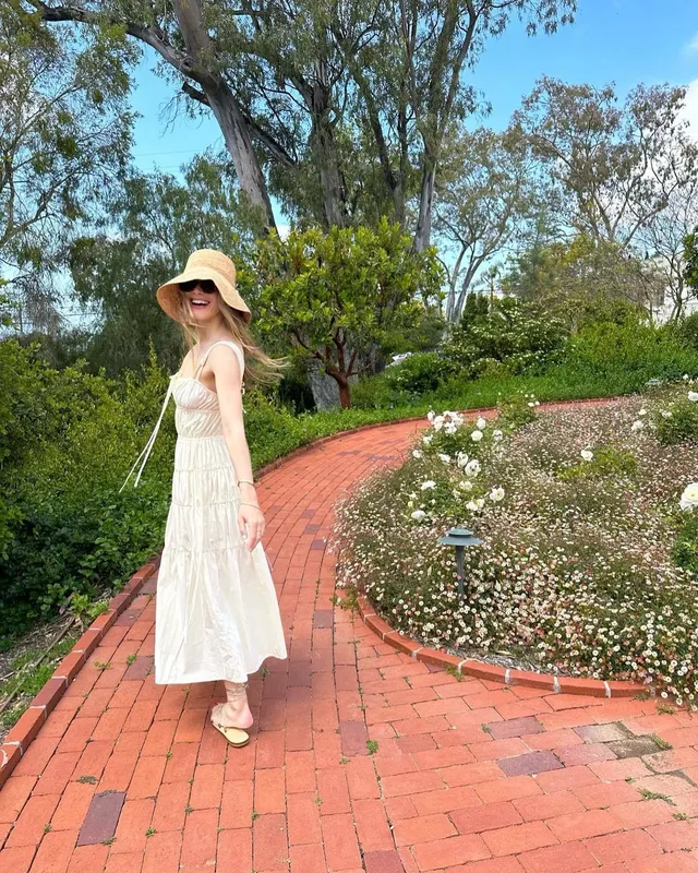 Halston Sage standing on a garden path in a white dress and light flat shoes.