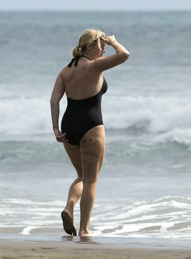 Kate Winslet walking barefoot by the sea in a black swimsuit.