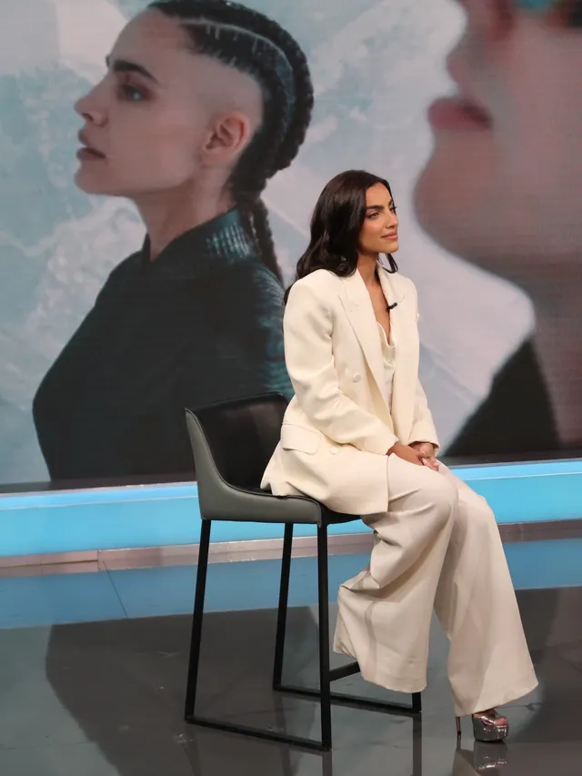 María Gabriela de Faría sitting onstage in a pale trouser suit and heels.