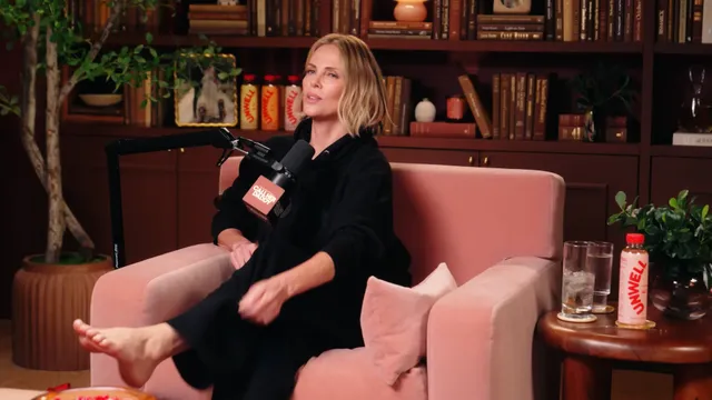 Charlize Theron seated on a pink talk show chair in a black outfit.