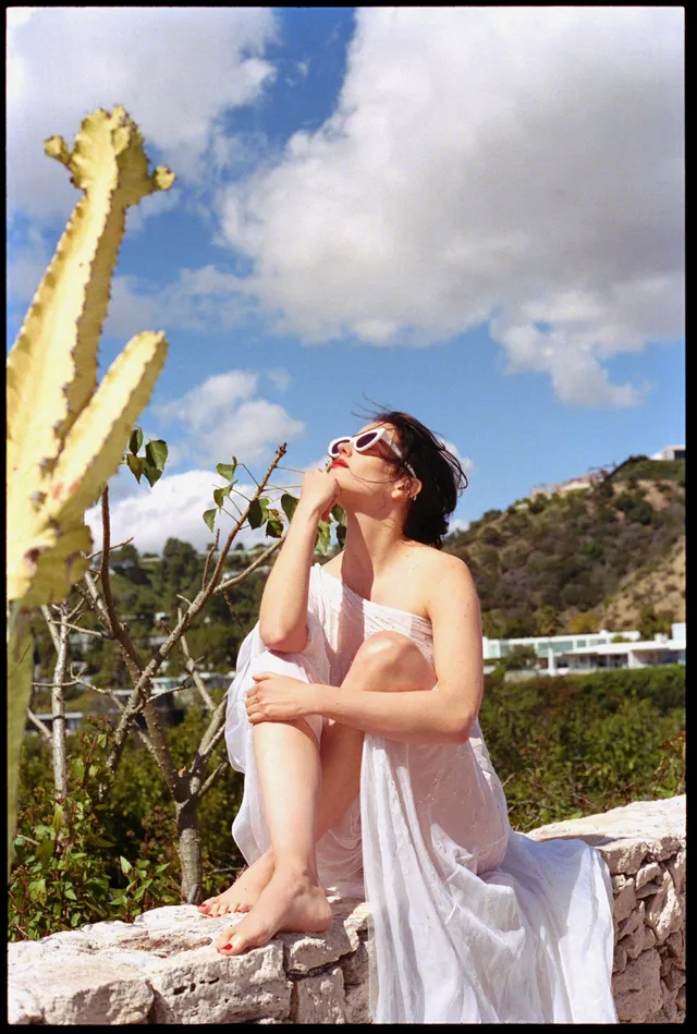 Eva Green seated outdoors beside tall cactus plants.