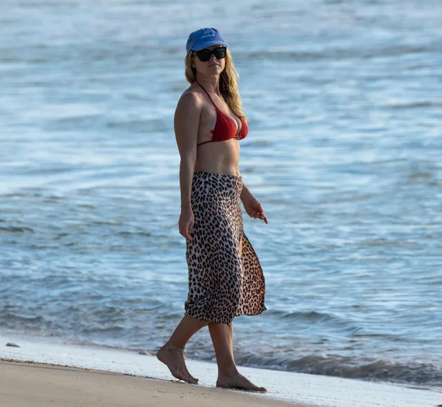 Alice Eve walking on a beach in a patterned wrap and blue cap.