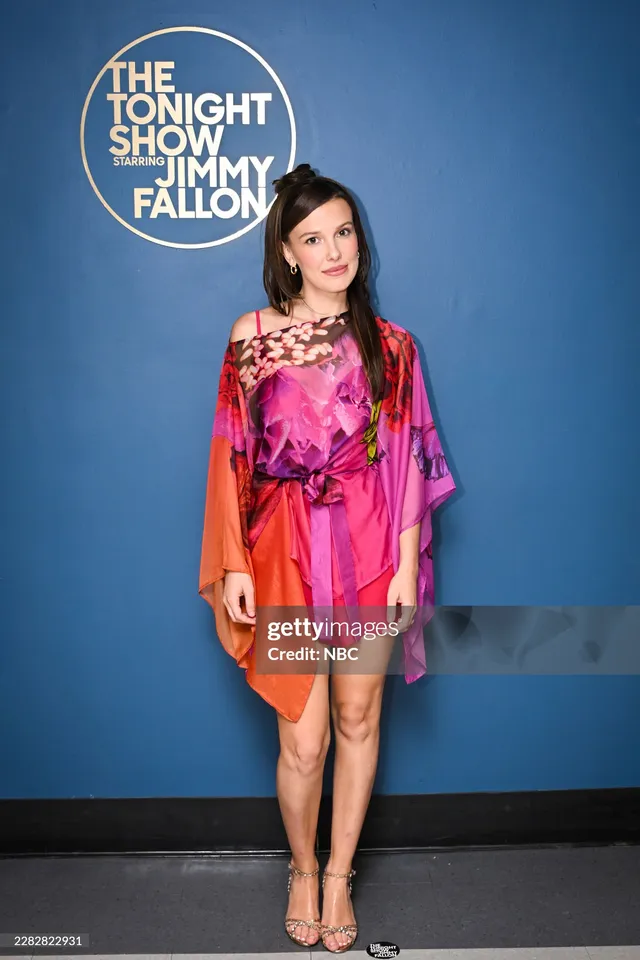 Millie Bobby Brown posing beside a television studio backdrop in a pink dress.