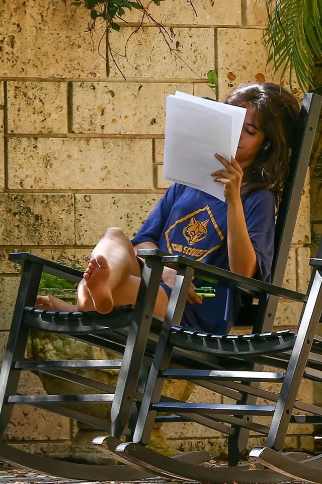 Camila Cabello reading in a chair outdoors with bare soles raised.