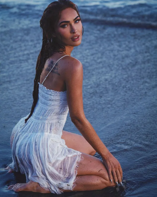 Megan Fox kneeling at the water's edge in a white dress.