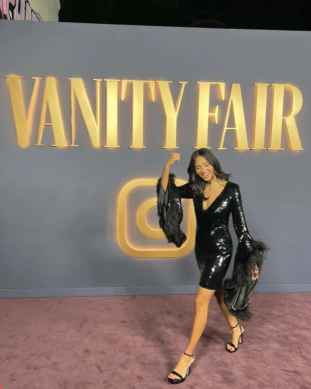 Greta Onieogou walking before an event backdrop in a black dress and heels.