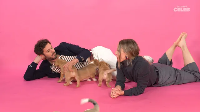 Kristen Bell lying barefoot on a pink floor beside another person and small dogs.