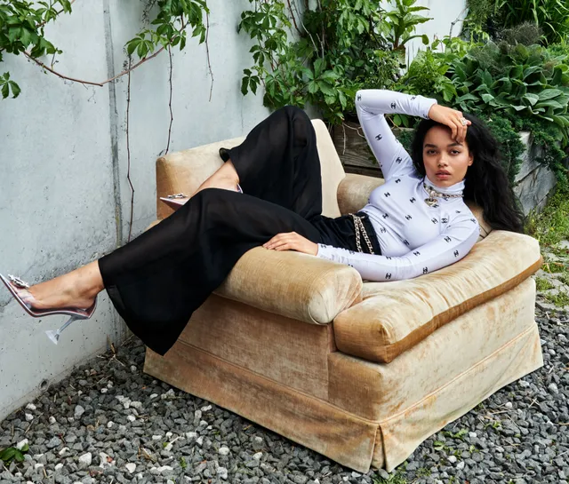 Whitney Peak reclining outdoors in an armchair in black trousers and metallic heels.