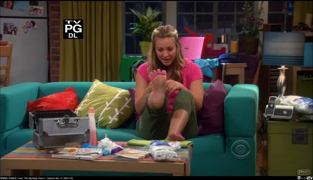 Kaley Cuoco holding a bare foot while sitting on a bright sofa in a television scene.