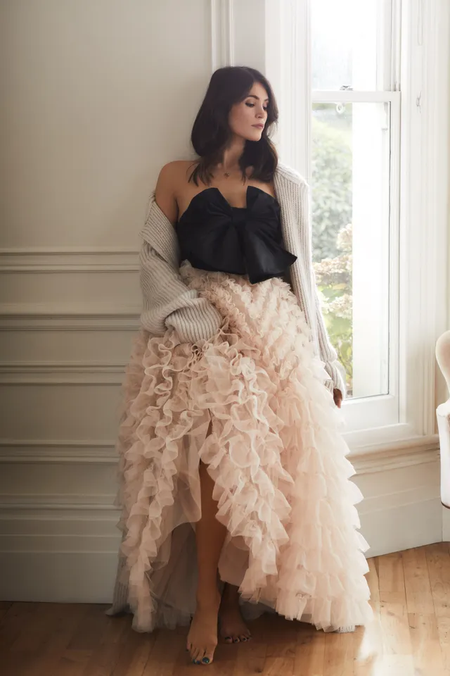 Gemma Arterton standing by a window in a layered pale skirt.