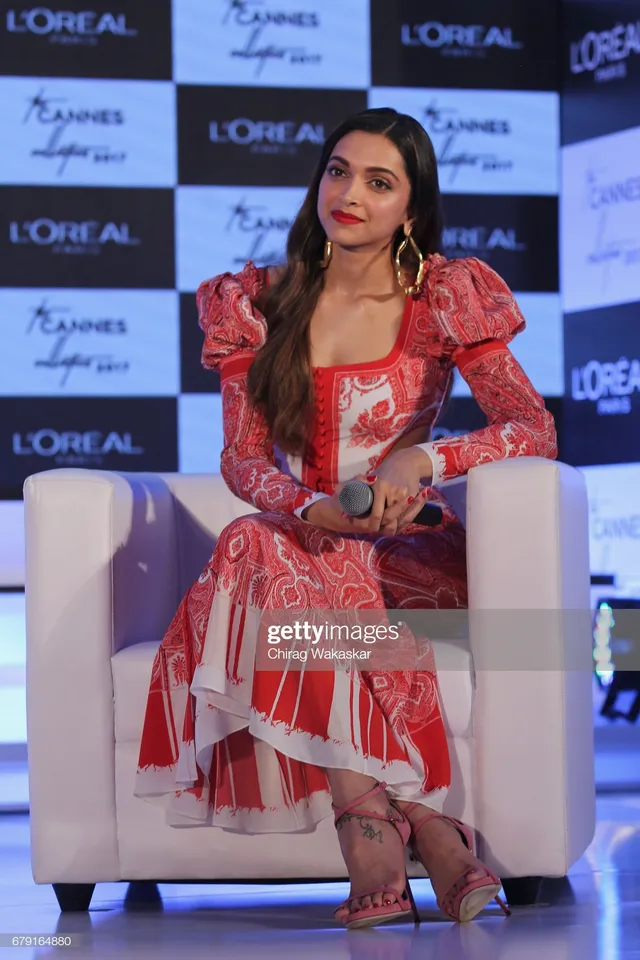 Deepika Padukone sitting in heeled sandals and a patterned dress at a promotional event.