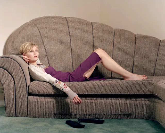 Kirsten Dunst lying barefoot on a couch in a pale top and purple trousers.