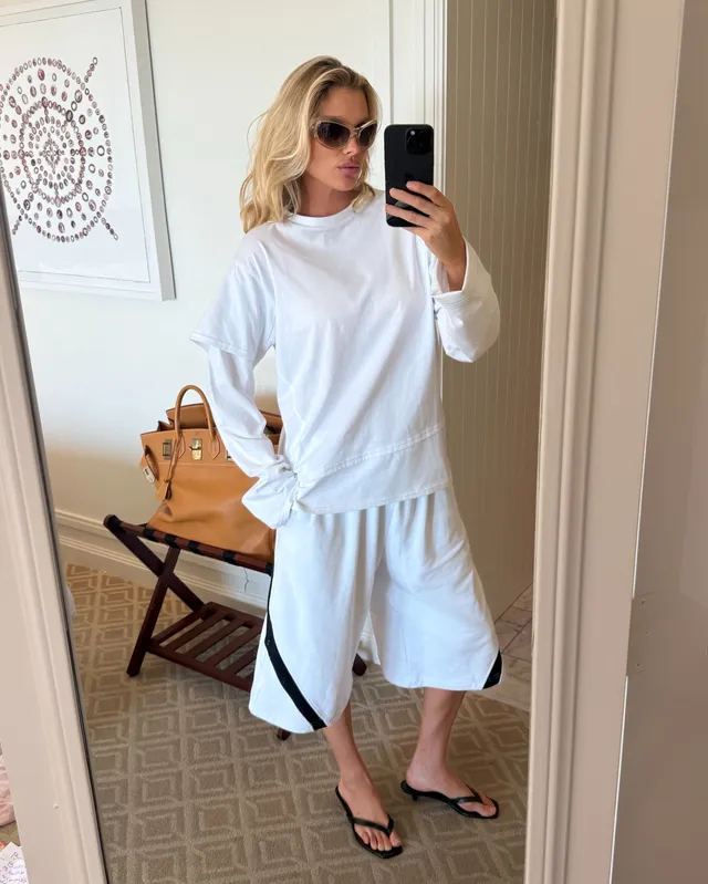 Elsa Hosk wearing flat sandals with a white sweatshirt and shorts in a mirror photograph.