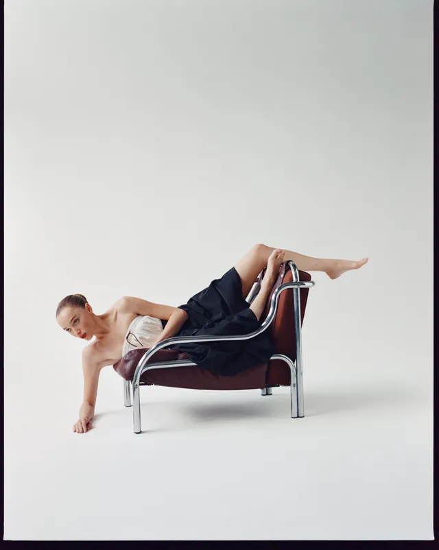 Phoebe Dynevor reclining across a sculptural chair in a studio.