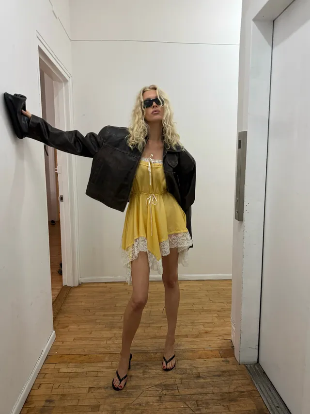 Elsa Hosk wearing black heeled sandals with a yellow dress and black jacket in a mirror photograph.