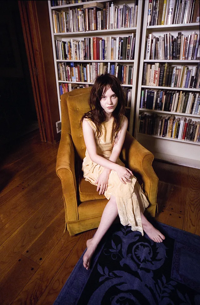 Sophie Thatcher sitting barefoot in an armchair in a yellow dress.