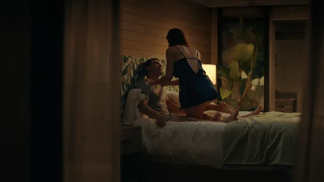 Alexandra Daddario seated barefoot on a bed in a softly lit screen scene.