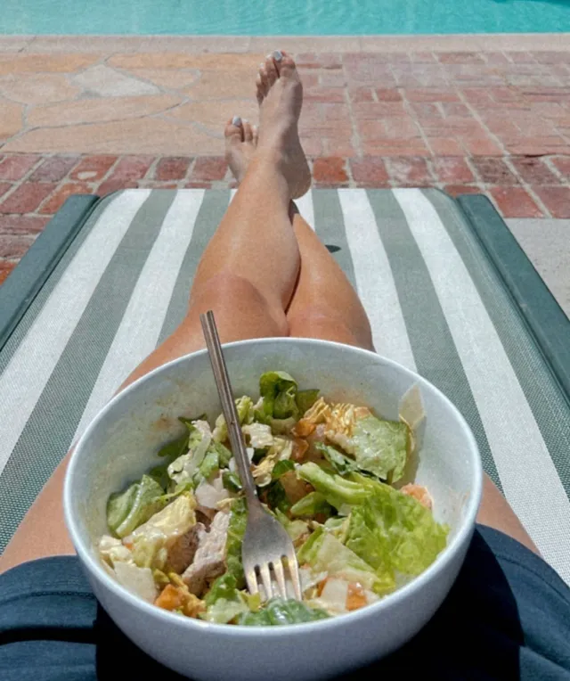 Brec Bassinger reclining barefoot on a pool lounger with a bowl of salad.