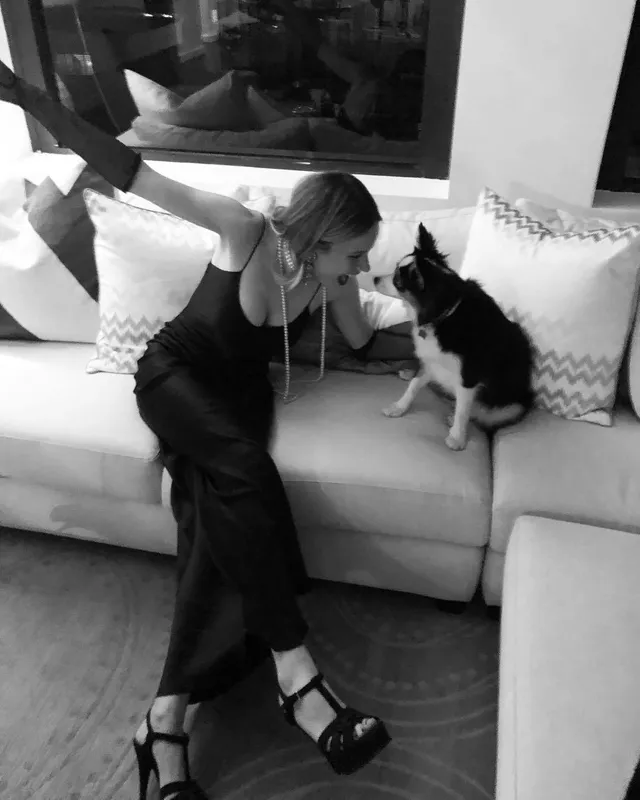 Halston Sage sitting on a sofa beside a dog in a black outfit and black heels.