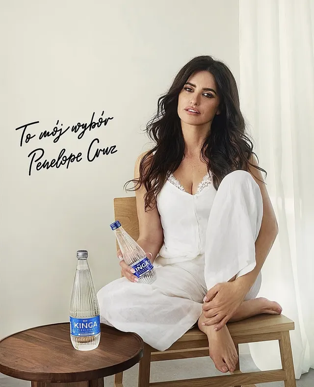Penélope Cruz seated in a white dress beside bottled water.