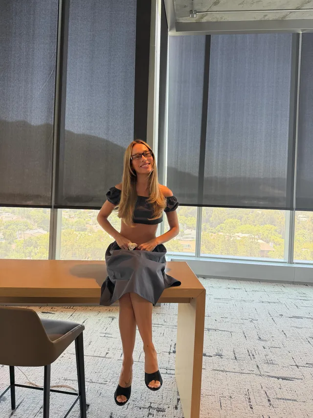 Ester Expósito sitting on a table wearing black heeled mules and a dark skirt during a screen scene.
