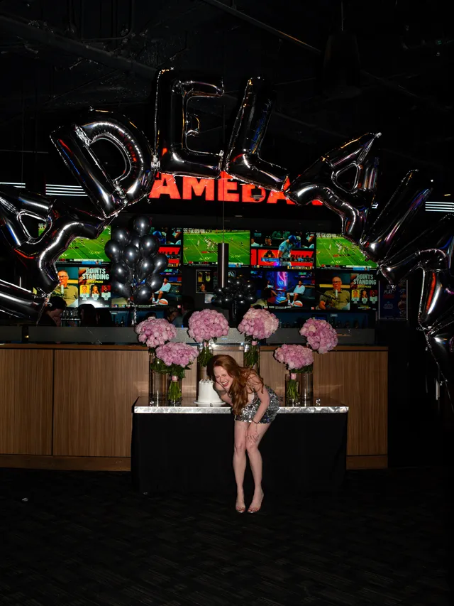 Madelaine Petsch sitting in a black outfit and heels beside event balloons.