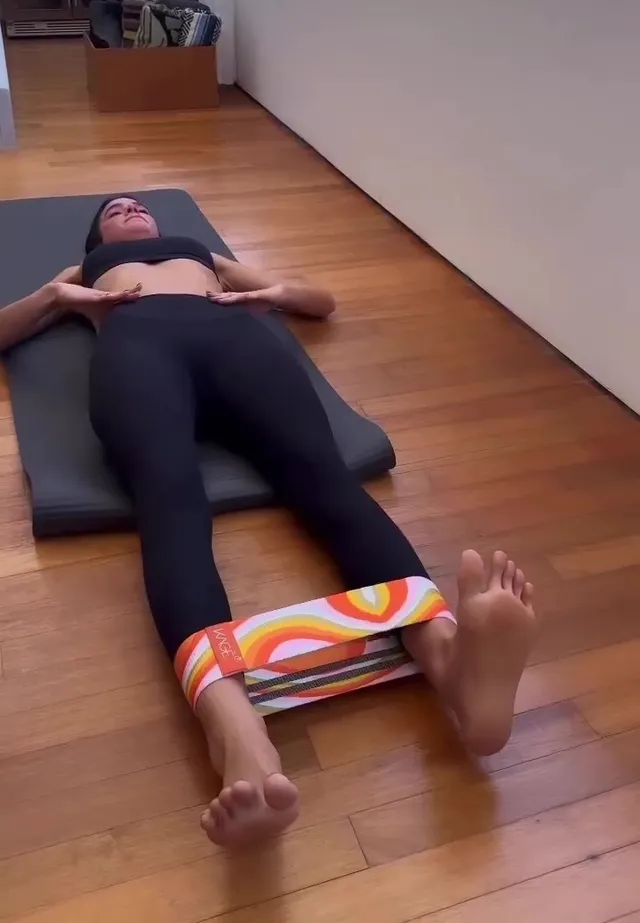 Bruna Marquezine lying barefoot on a gym mat with a resistance band around her legs.