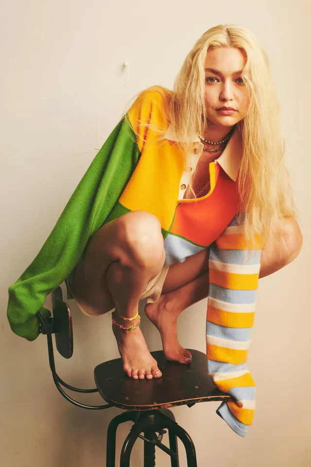 Gigi Hadid crouching barefoot on a stool in a colorful outfit.