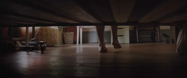 Chloë Grace Moretz standing barefoot on a wooden floor beside a bed.