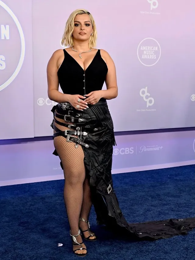 Bebe Rexha standing at an event in a black gown and metallic heels.
