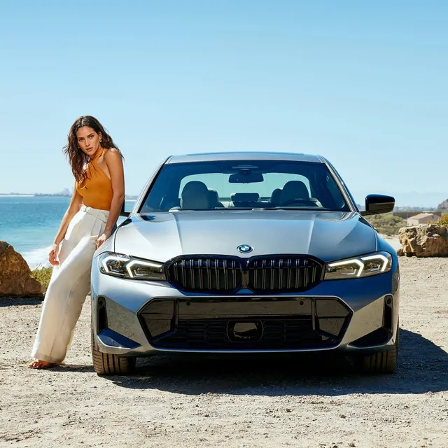 Adria Arjona standing beside a car in an orange top and white trousers.