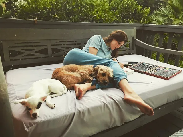 Alexandra Daddario relaxing barefoot on an outdoor sofa with dogs.