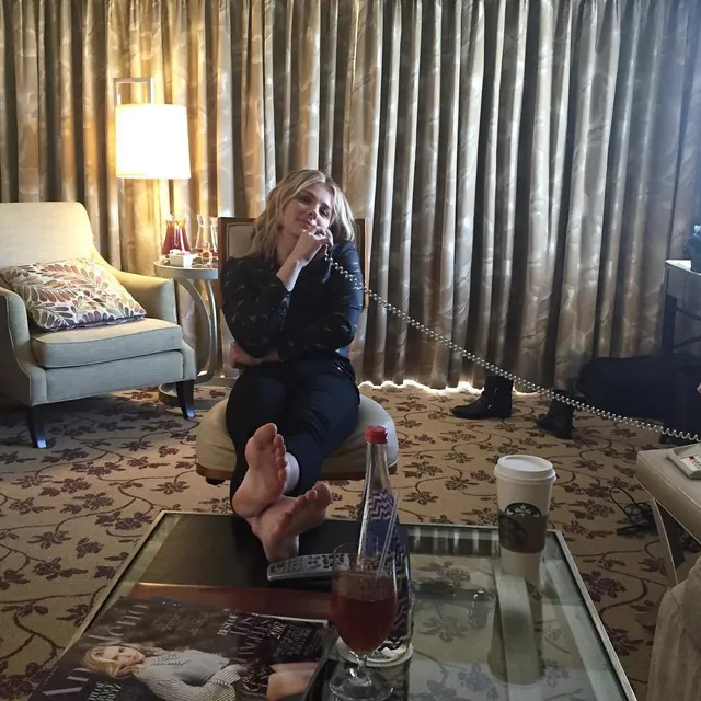 Chloë Grace Moretz sitting indoors with bare soles resting on a glass table.